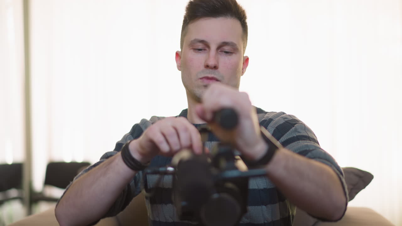 Man adjusting video camera setup with focus on handheld equipment, preparing for filming. Modern home studio with green walls and natural light in background, setting up for content creation