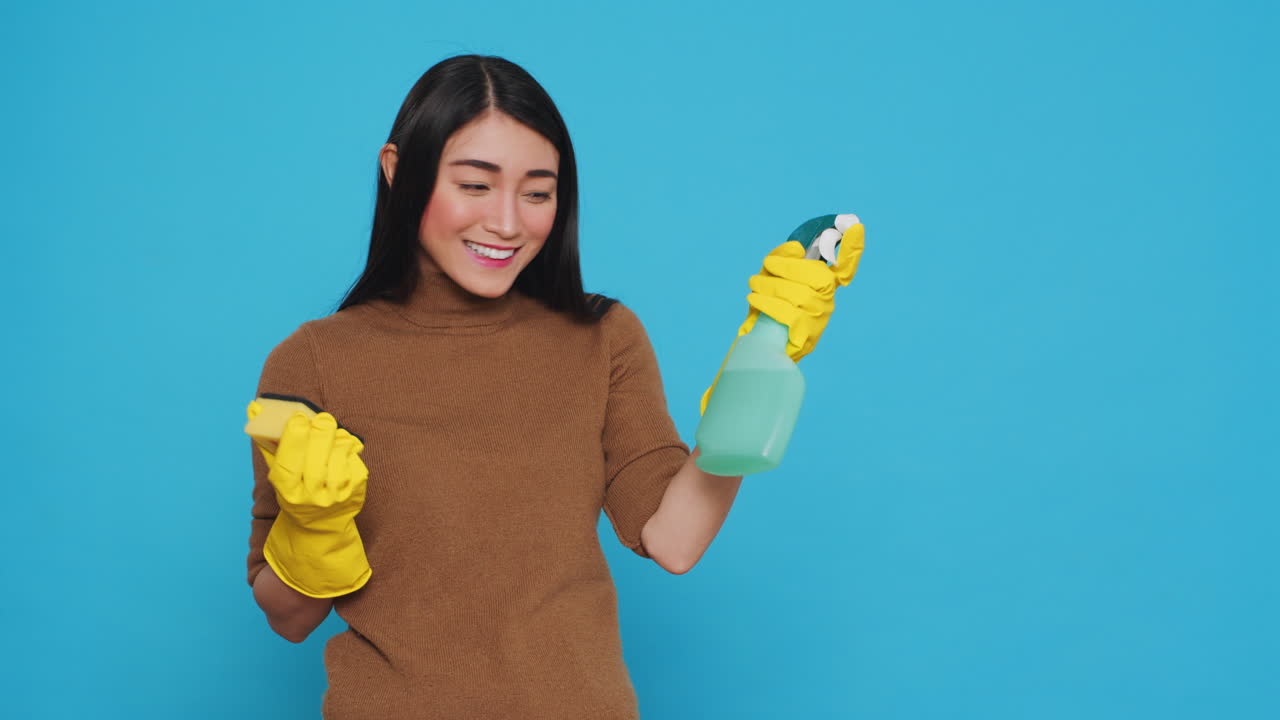 Cheerful asian housewife wearing rubber gloves while cleaning house using sponge and detergent spray