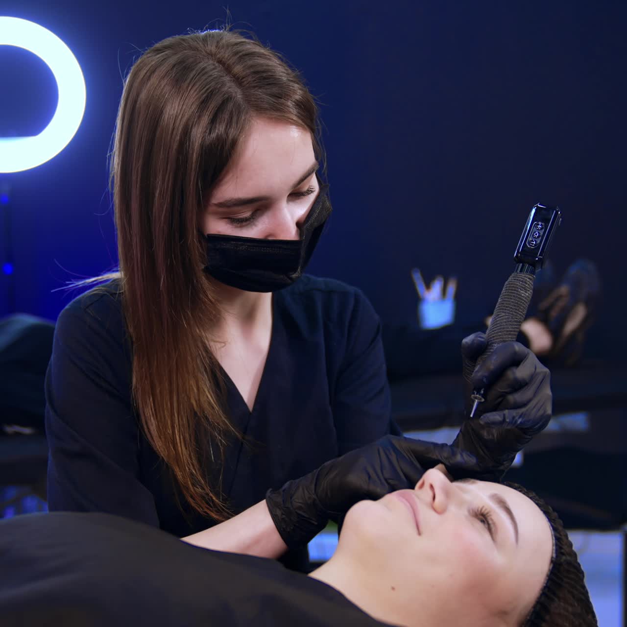 Long-haired brunette brow master uses special machine for brow make up. Specialist sits over the client lying on the couch in salon