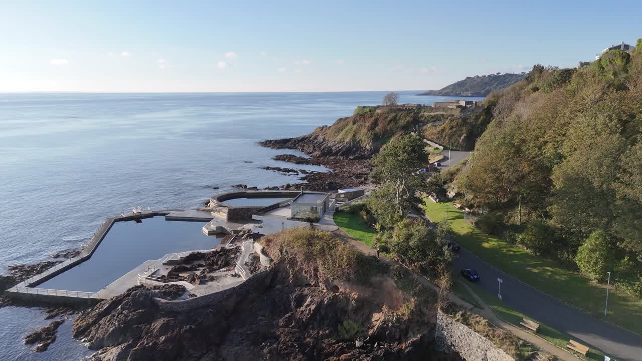 Forward drone flight over the seawater bathing pools at Havelet Bay Guernsey on calm day in late afternoon sunshine close in to wooded cliffs and Clarence Battery in the background