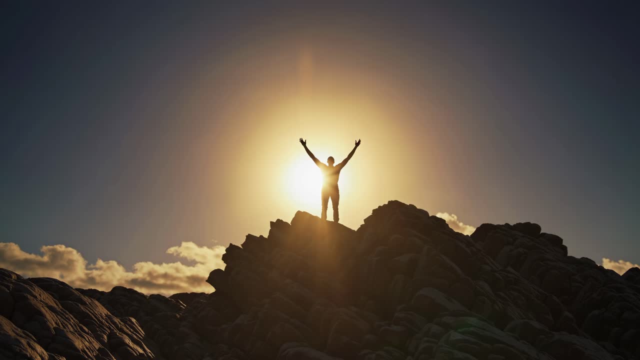 Silhouette di una persona con le braccia alzate su una cima rocciosa al tramonto, ripresa da una angolazione dal basso