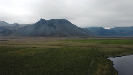 Umlaufende-Drohnenaufnahme-Isländischer-Berge,-Felder-Und-Eines-Sees-Mit-Dramatischem-Nebel-Im-Hintergrund
