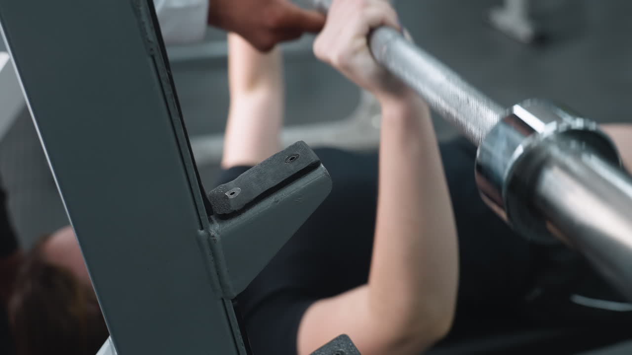 close up of woman gripping barbell during bench press as male trainer assists in lifting, highlighting hands, metal bar, and supportive guidance in focused gym environment with equipment visible