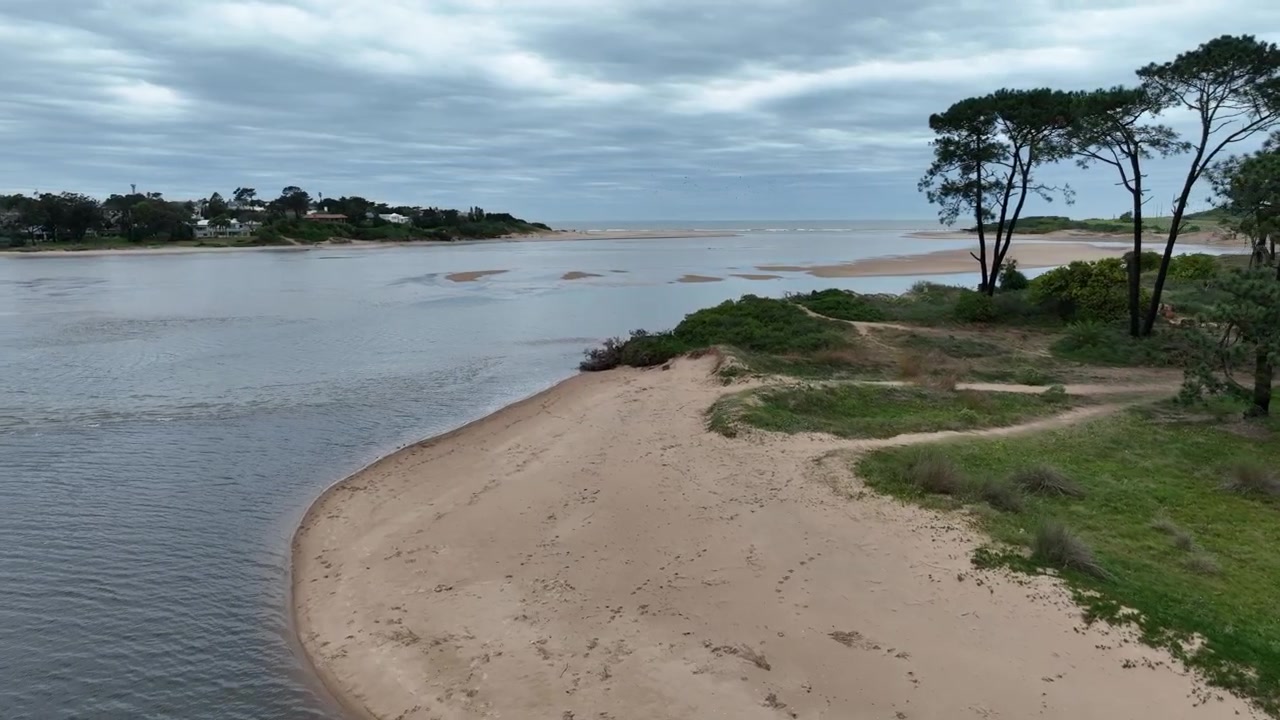 Arroyo Maldonado forward push along a sandy river bend with green vegetation and distant houses in Punta del Este