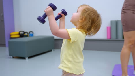 adorable little girl holds dumbbells, playfully mimicking workout routine in bright fitness room, smiling child enjoys lifting small weights while adult figure stands nearby