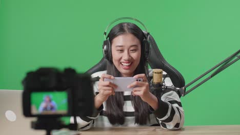 a smiling asian woman gamer with headphone and computer playing mobile phone game while shooting video by camera on the green screen