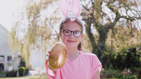 portrait of girl wearing bunny ears holding chocolate egg on easter egg hunt in garden