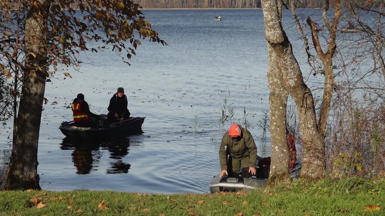 Fishermen in a boat on a blue lake, casting lines and catching fish on a calm day. The water reflects the autumn landscape, enhancing the serene and seasonal feel.