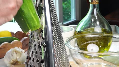 grating a cucumber. making tzatziki greek or mediterranean sauce or dip for a party. dip or sauce in a green bowl on a wooden cutting board with dill, green napkin and a grater.