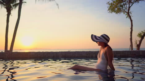 a young woman, in a one-piece bathing suit and floppy sun hat, relaxes in the shallow water of a resort pool at sunset