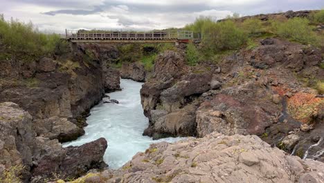 view over the bridge located over the ligthblue waterfall barnafossar with alot of rocks and green bushes surrounding the water