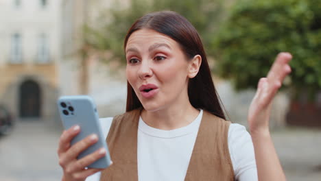 excited european young woman using smartphone typing browsing celebrating win on urban city street