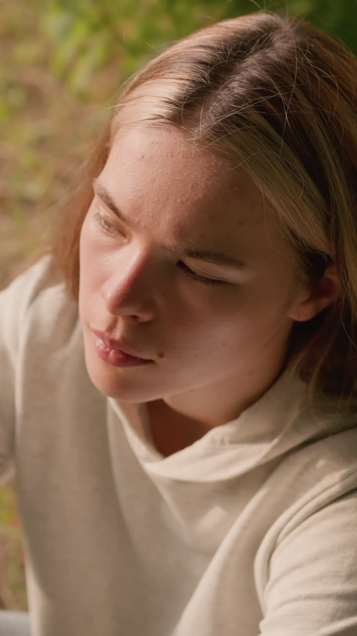 joven sentada en el suelo bajo un árbol con la mano sobre la rodilla, centrándose atentamente en algo, el follaje seco esparcido alrededor añade a la configuración natural