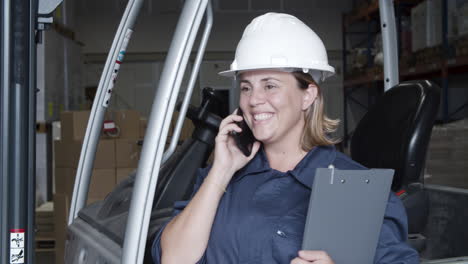 happy caucasian female worker talking on phone, standing in storehouse and holding a clipboard