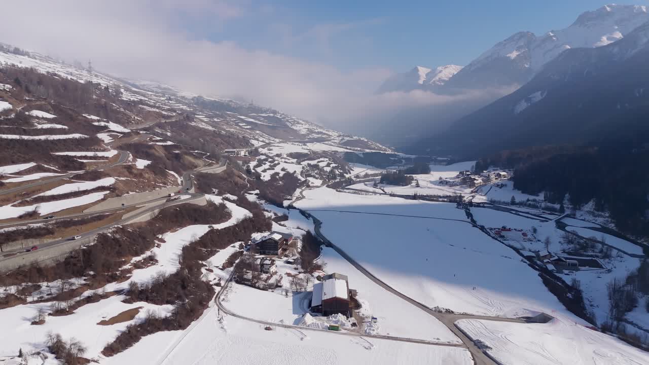 Swiss Highway with traffic on hillside of alps mountains. Sunny day with snowy fields and mountains in Scuol, Switzerland. Aerial forward wide shot.