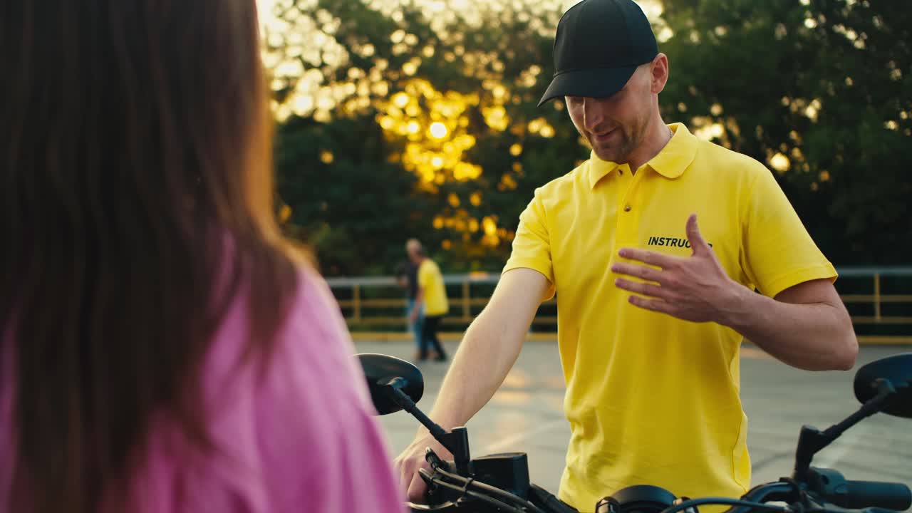 A guy in a cap in a yellow t-shirt of an instructor sits on a motorcycle and shows a girl in a pink t-shirt How to sit on a motorcycle. driving school instructor