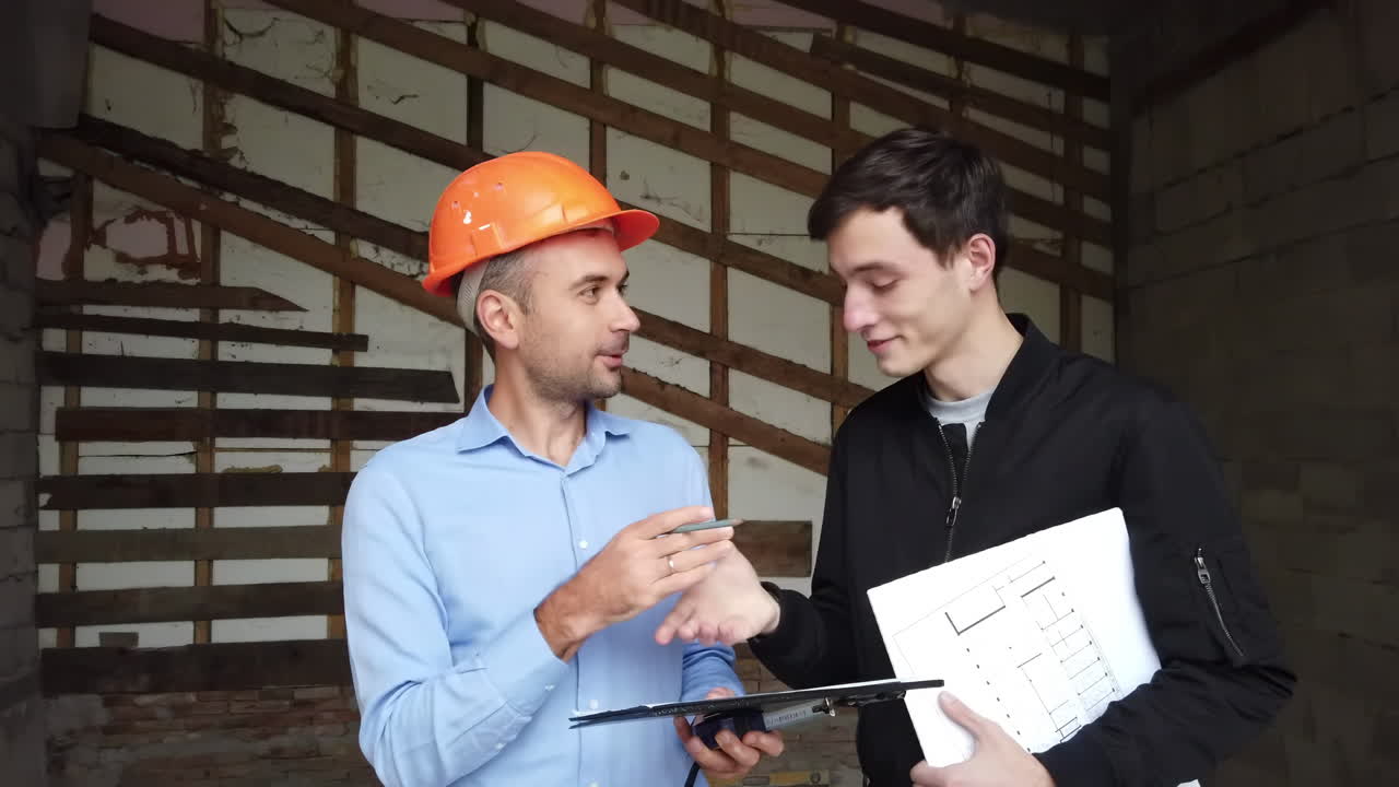 A site manager wearing an orange safety helmet explaining plans to a colleague on a construction site