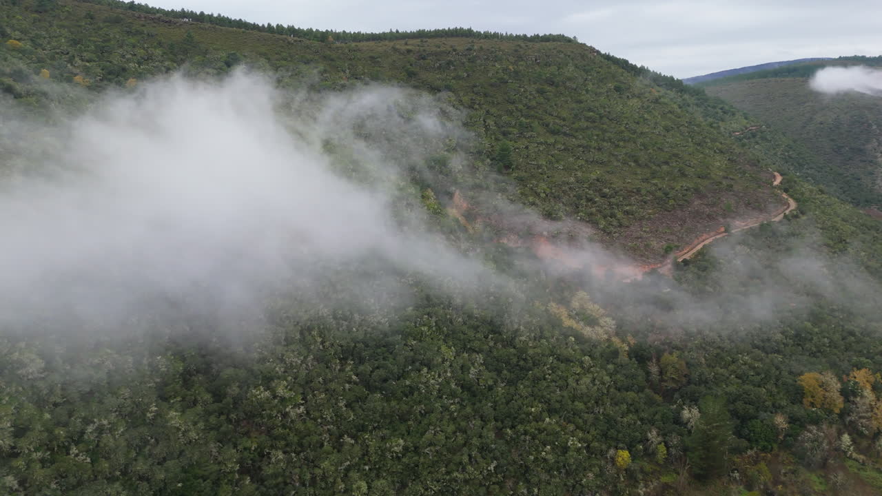 Aerial drone view of mist drifting across lush green hills with a winding dirt road cutting through the mountain landscape on a cloudy day