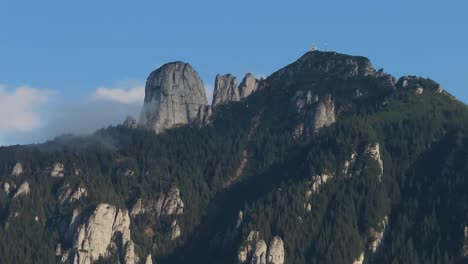 Aerial-close-up-shot-showing-tall-mountains-peaks-covered-in-dense-coniferous-green-forests,-blue-sky