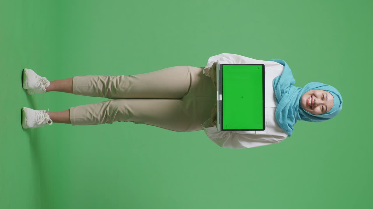 Full Body Of Asian Muslim Woman Smiling And Showing Green Screen Laptop To Camera While Standing In The Green Screen Background Studio