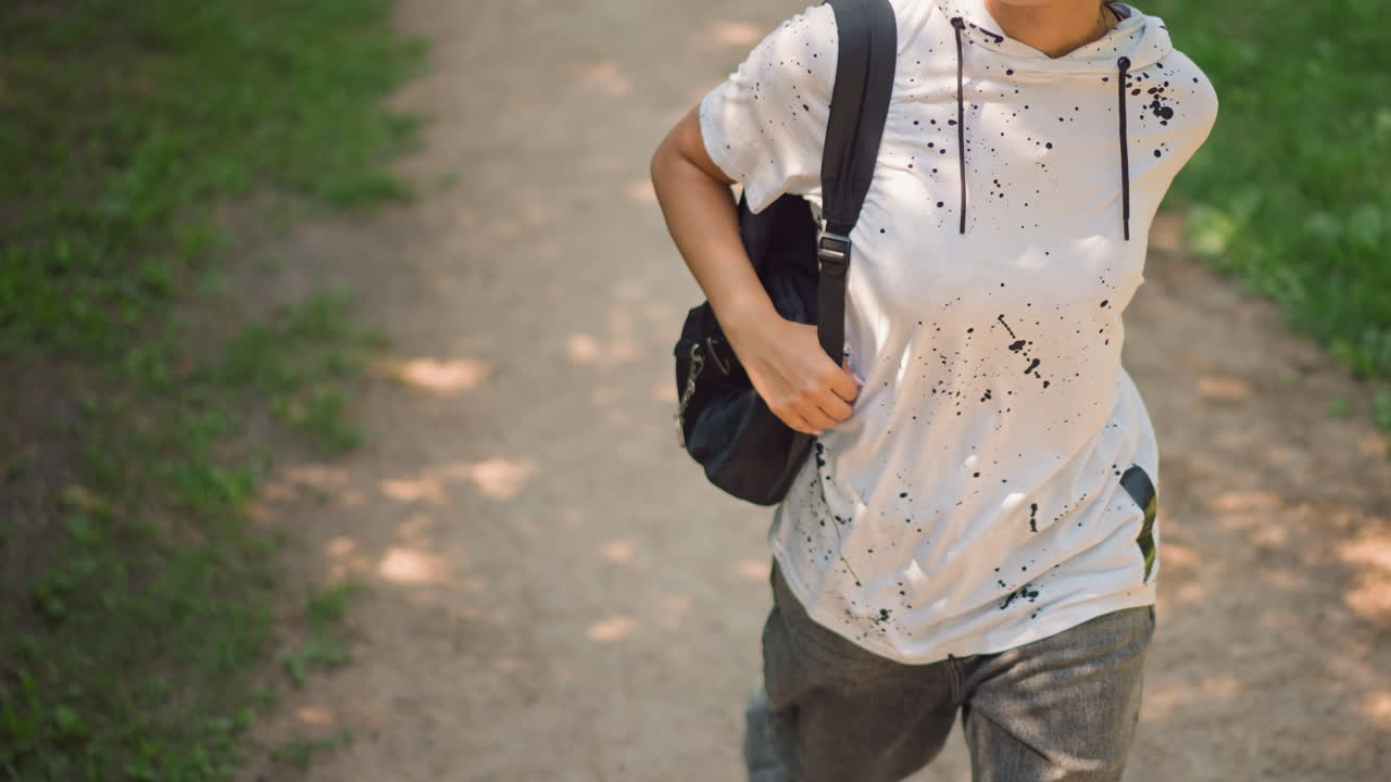 나뭇잎이 깔린 길에서 백팩 끈을 맨 캐주얼한 복장의 사람이 걷고 있습니다. 앞모습 토르소 샷에서는 손목에 스마트워치가 보입니다. 흰색 반점이 있는 후드티를 입고 안정적으로 걷다가, 얼룩덜룩한 햇빛이 비치는 곳을 지나면서 끈을 조절하고 주머니에 손을 넣습니다.