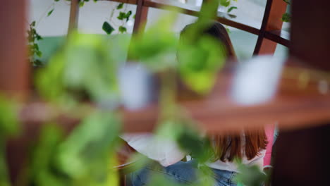 close-up of wooden frame decorated with green leaves, partially revealing a woman seated, using her phone, with a shopping bag beside her in a relaxed and stylish indoor setting