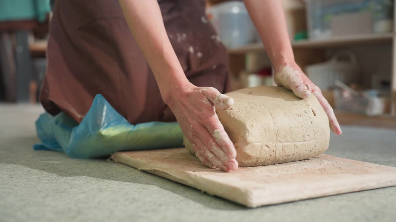 Kneeling white potter lifts clay block onto board, preparing slab for throwing, workshop shelves and tools in background, practical motion and press, hands shaping and positioning, instructional