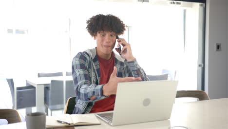 un joven biracial está usando una computadora portátil y hablando por teléfono en casa en la cocina