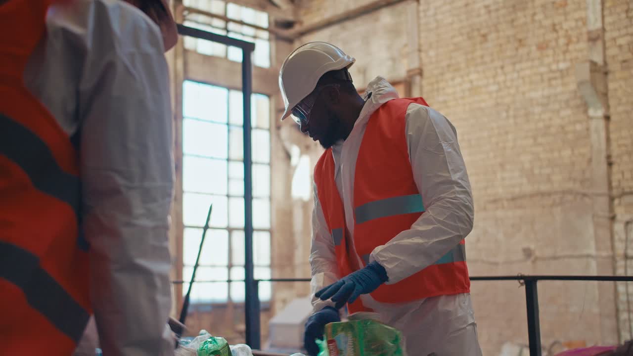 Shooting close up a man with Black skin in a special white protective uniform together with his colleague sorts and sorts garbage at a large waste processing plant near a conveyor belt