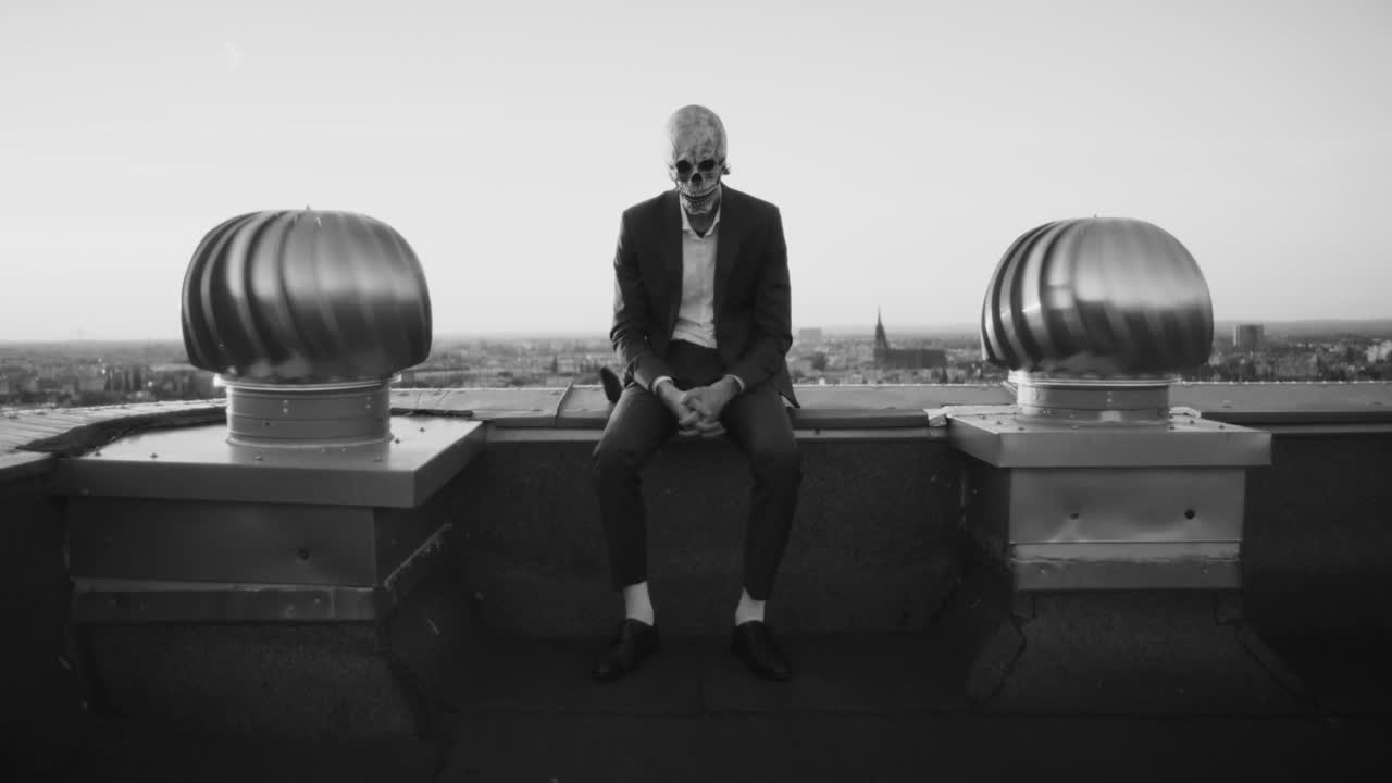 A person in a suit and skull mask sits alone on a city rooftop