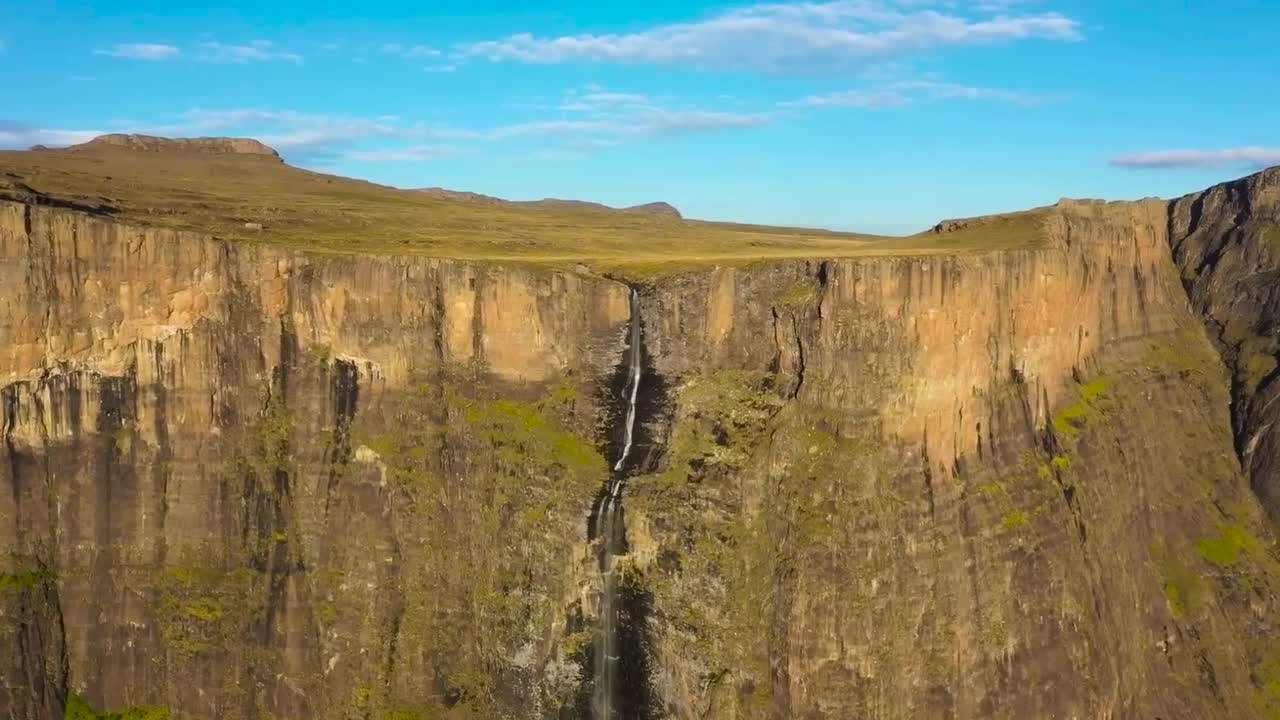 Aerial drone footage flying closer to Dragon's Mountains of Royal Natal National Park Tugela Falls Waterfall that is falling off a steep tall brown cliff in South Africa during sunny day, green land.