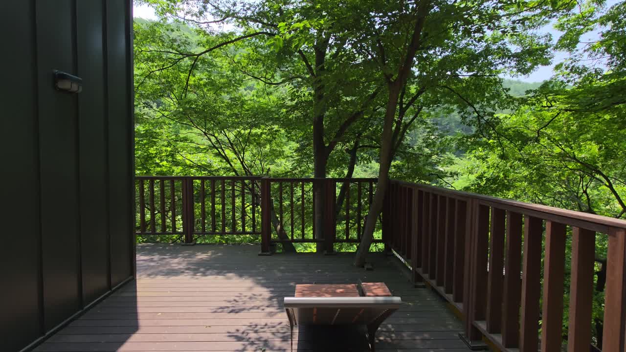 Camera glides forward along a wooden terrace beside a modern cabin, moving through dappled sunlight and lush green trees, drawing viewers into the tranquil forest setting of Maninsan Ecological Park