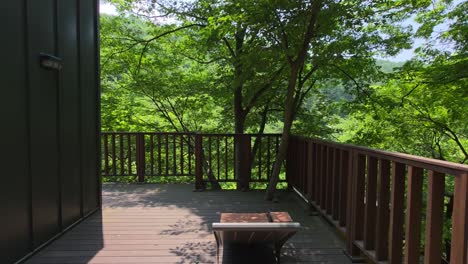 camera glides forward along a wooden terrace beside a modern cabin, moving through dappled sunlight and lush green trees, drawing viewers into the tranquil forest setting of maninsan ecological park
