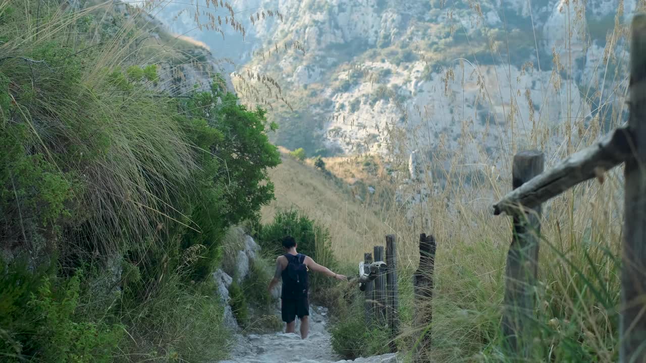 Downward Tracking Wide Shot of Young Asian Adult Male Walking Down Dry Stony Path in Beautiful Mountain Valley
