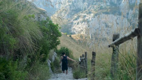 downward tracking wide shot of young asian adult male walking down dry stony path in beautiful mountain valley