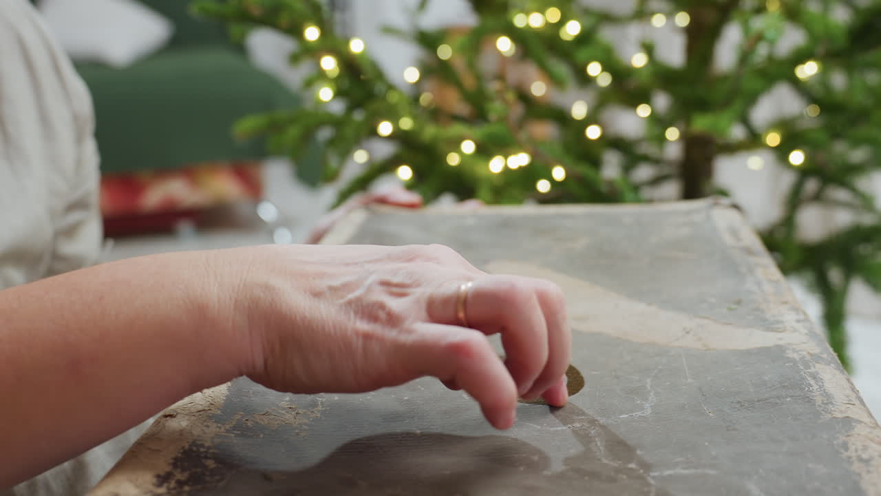Hand view of woman drawing playful shapes with wet finger on dusty surface, capturing tactile creative moment with blurred glowing Christmas tree in background, evoking cozy festive indoor holiday