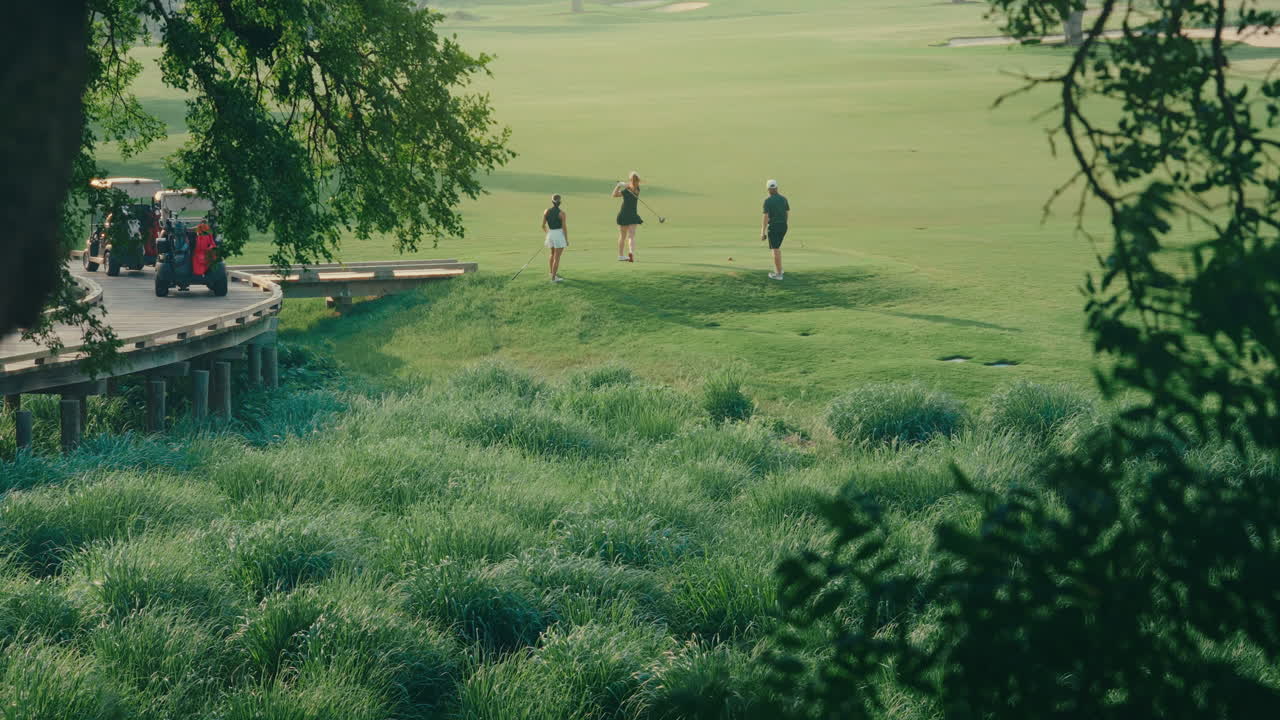Distant shot through trees capturing one male and two female golfers on the tee box preparing to tee off. Scenic golf course setting with natural framing, bright weather, and a calm outdoor atmosphere