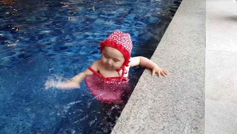 playful baby girl in bathing suit stand on swimming pool step playing and slapping water by her hand