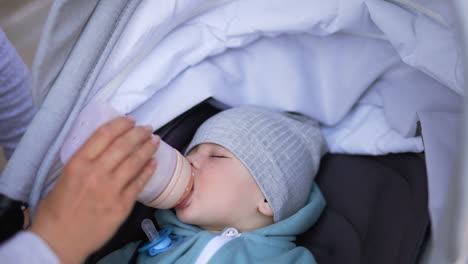 sleepy adorable baby being fed from bottle during walk. caucasian child lies in stroller falling asleep and suckling bottle. close up.