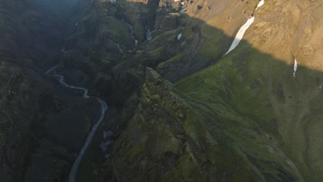 drone shot of a deep canyon in iceland, showcasing steep cliffs, a winding river, and rugged terrain with patches of snow