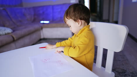 two-year-old baby boy sitting at desk looking at his picture. little kid drawing at home.
