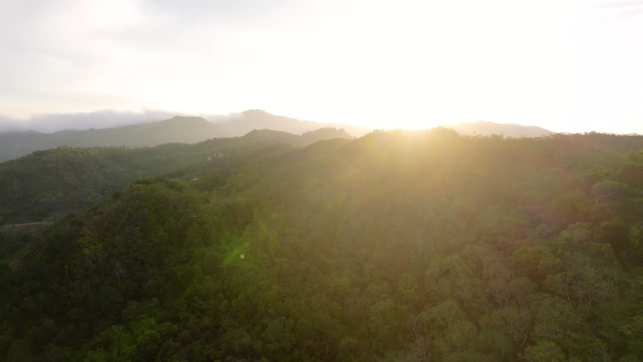 Golden sunlight peaks behind lush tropical rainforest mountains, aerial