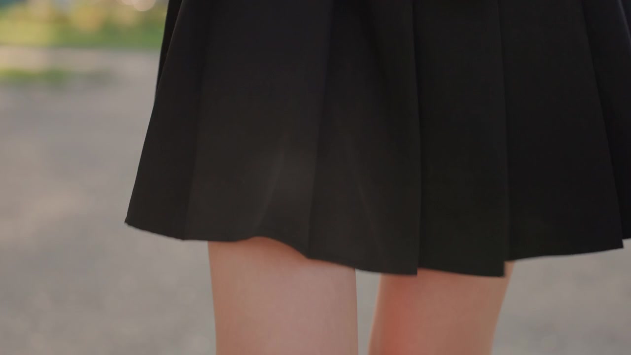 黒いスカートを着用し、赤いバッグを持った若い女性が屋外を歩くクローズアップ。柔らかな日差しの下、緑豊かな小道を歩く女性の手は自然に動いている