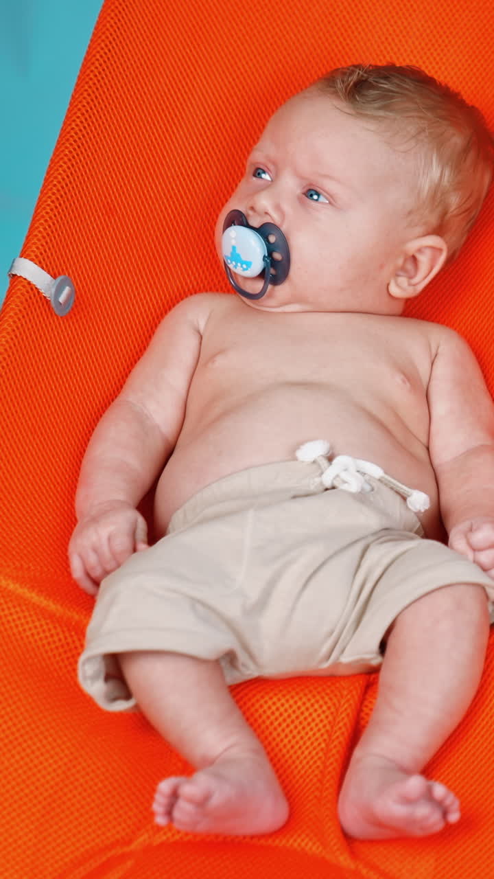 Adorable Caucasian infant relaxing on the imitated beach. Lovely kid in shorts and with pacifier looking attentively aside. Vertical video