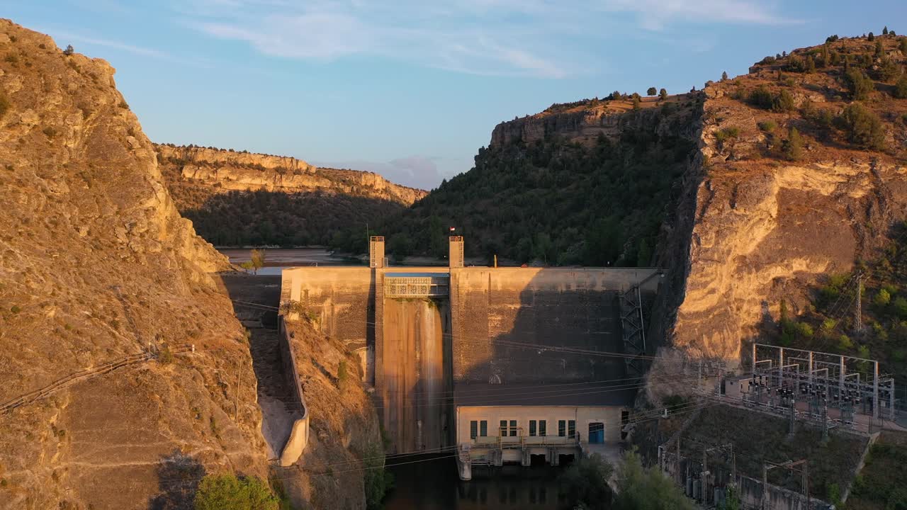vuelo de avión no tripulado ascendiendo frente a una planta hidroeléctrica en una presa de agua construida en los bordes de un cañón de piedra naranja con formaciones forestales de un río en la hora dorada en segovia, españa