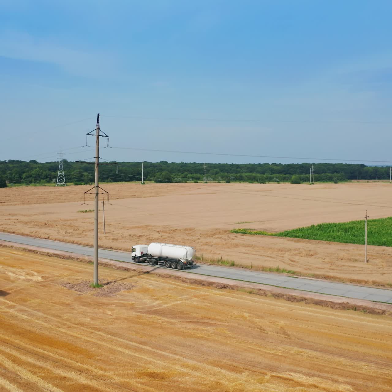 Road crossing the agricultural farmlands and trucks going by it. Tractor loaded with hay bales rides by the cut field. Top view