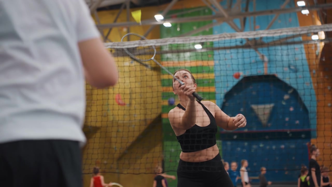 Atleta mulher devolvendo o volante durante uma intensa troca de pontos de badminton em quadra coberta com o parceiro do outro lado e uma parede de escalada ao fundo, reações expressivas, voleios ritmados e smashes de ataque