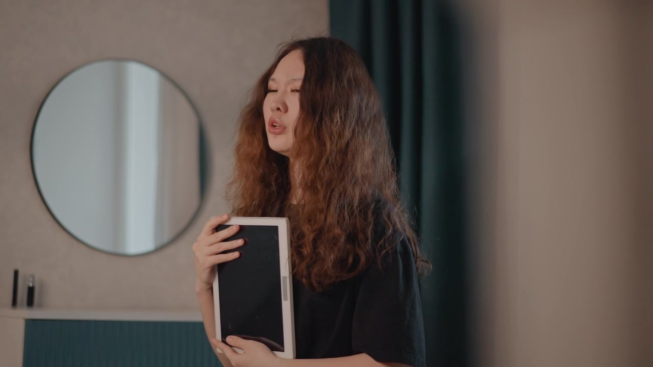 Giovane donna tiene un tablet al petto mentre parla e gira la testa accanto a una parete a pannelli color verde acqua con uno specchio rotondo, creando una scena interna rilassata con gesti espressivi, luce soffusa e un'atmosfera colloquiale.
