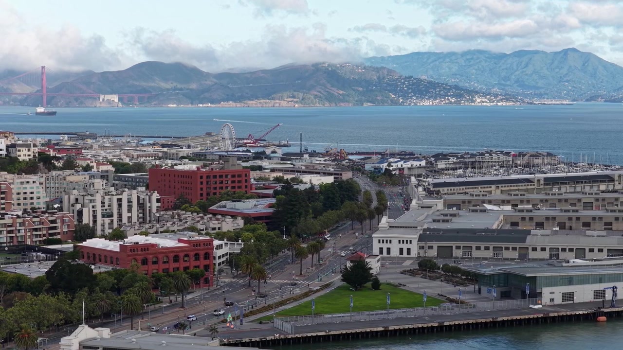 Una vista aerea cattura il lungomare di San Francisco, l'iconico Golden Gate Bridge e la baia panoramica sotto la morbida luce del mattino. Ampie riprese aeree con un vivace paesaggio urbano, acque calme e un cielo striato di nuvole.