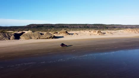 drone view of offroad 4 wheel drive red car with rooftop tent driving on sand on a long beach on a sunny day at 90 miles beach, northland, new zealand.